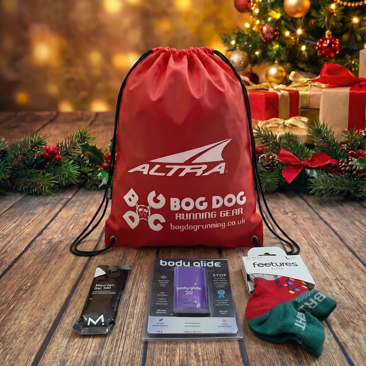 Bronze runner’s Christmas gift set: Feetures Elite Christmas socks, Body Glide Foot Glide and a Maurten Gel 100 in front of a red Bog Dog and Altra drawstring bag